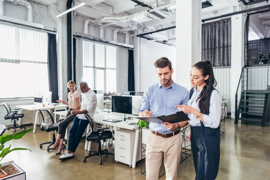 Two people looking at a folder in an office while two other people talk in the background.