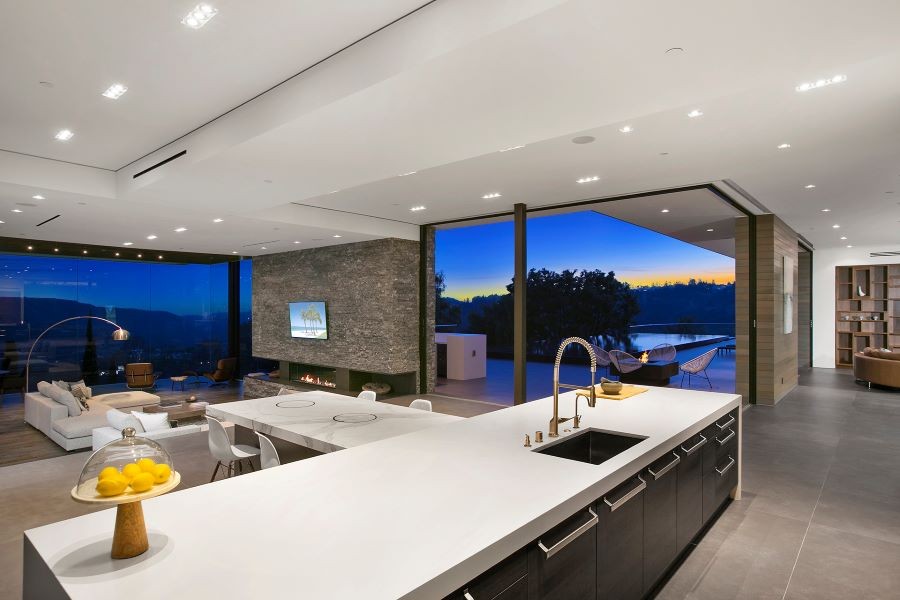 An open floor plan depicting the kitchen and living area with a TV above the lit fireplace and in-ceiling speakers.