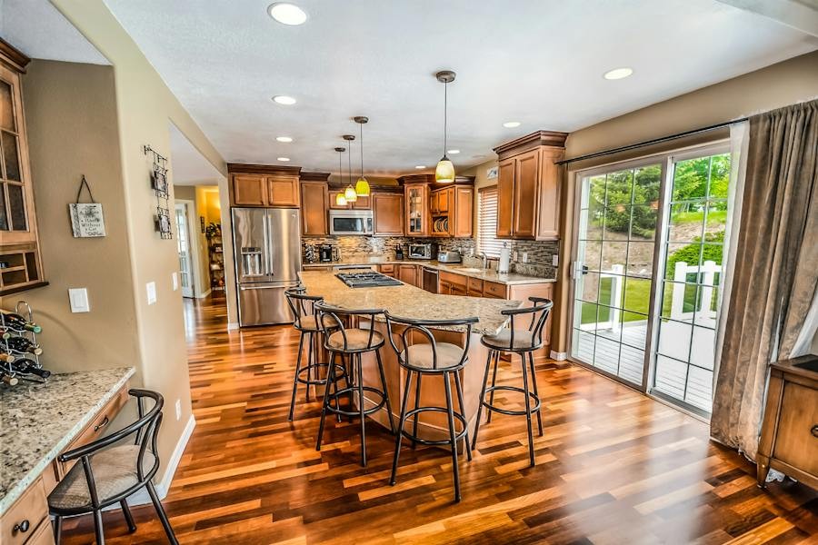 An updated kitchen with hardwood floors and granite countertops, recessed lighting and fixtures hanging above the island, a sliding glass door to the right. 