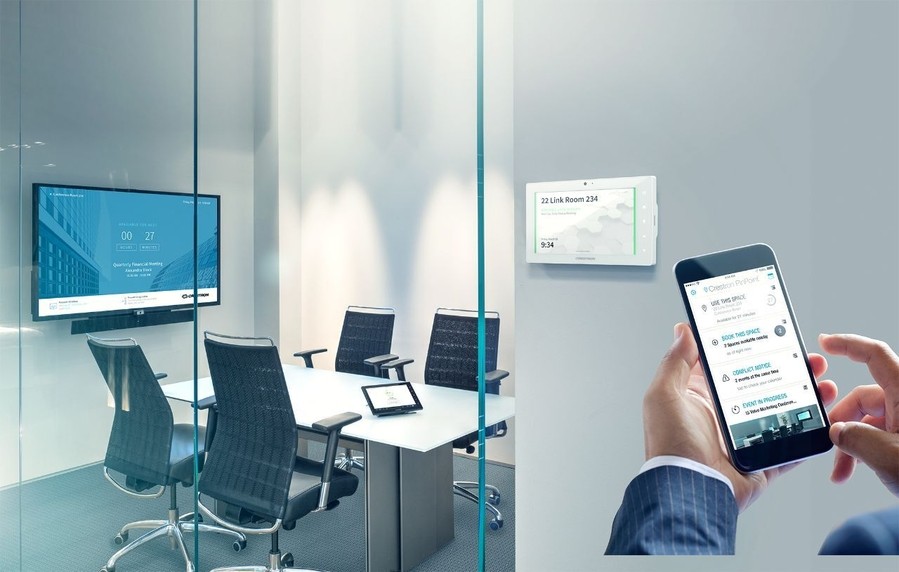 A person standing outside a video conference room with a room scheduling panel, pulling up an app on their smartphone. 