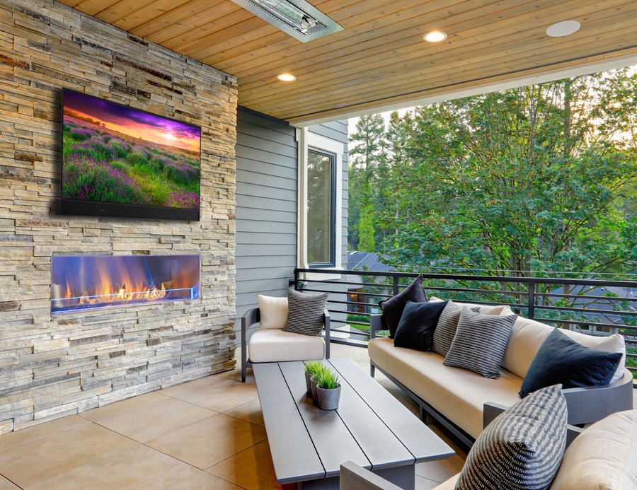A home’s patio with a couch, in-ceiling speakers, and a Séura outdoor TV above a fire features.\